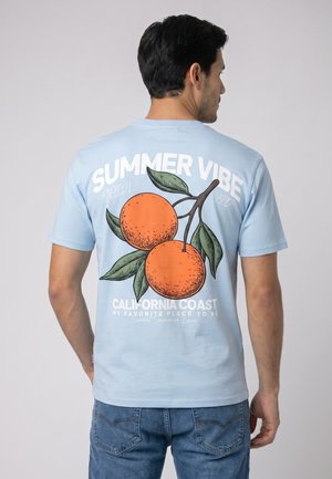 Man wearing a light blue t-shirt with two illustrated oranges and the text "Summer Vibe California Coast" on the back.