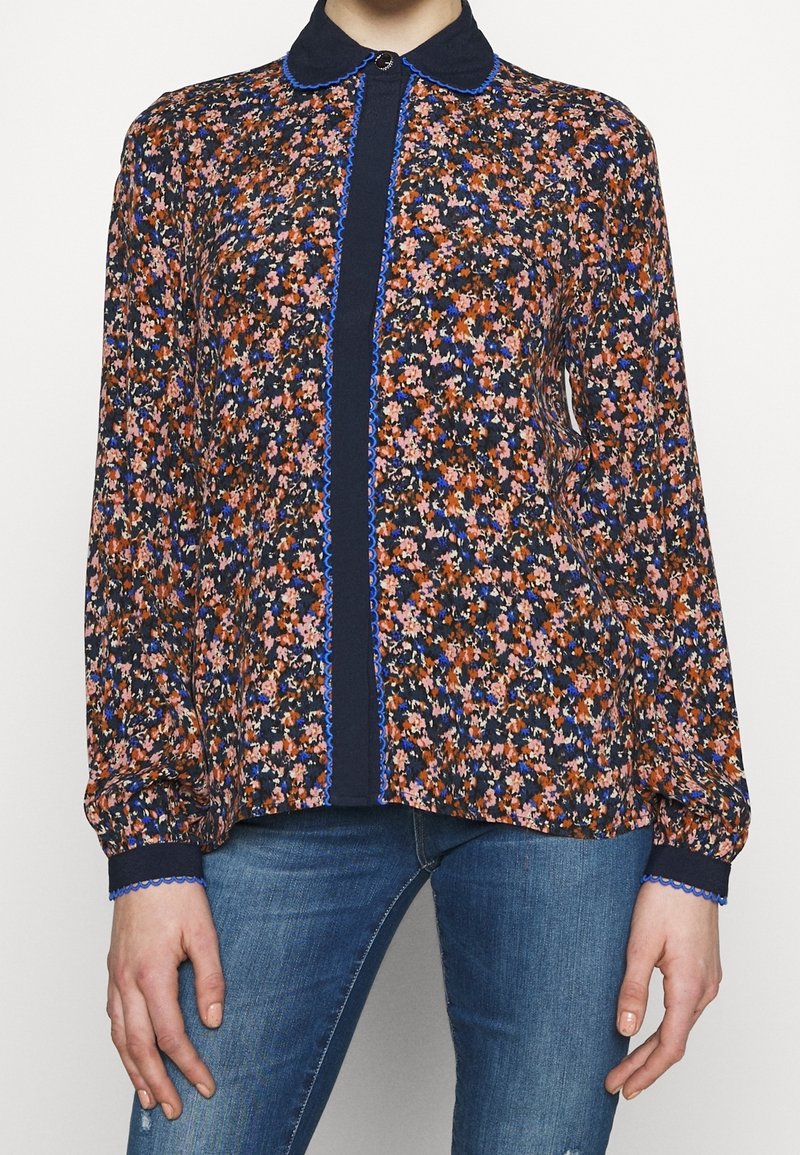 Femme portant un chemisier fleuri à manches longues avec une bordure bleue et un col bleu foncé, associé à un jean bleu.