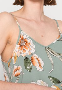 Top in seta floreale verde con fiori arancioni e bianchi. Caratterizzato da spalline sottili, scollatura a V e tessuto leggero con una consistenza morbida.