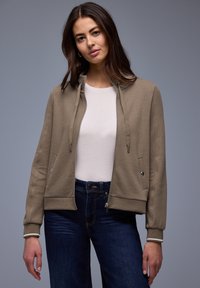 Joven con cabello largo y oscuro, vestida con una chaqueta beige con cremallera sobre una camiseta blanca y jeans azul oscuro, de pie frente a un fondo gris.