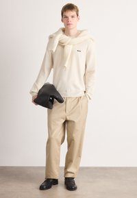 Giovane uomo con un maglione beige con logo "BOSS", sciarpa a maglia abbinata sulle spalle, pantaloni beige, scarpe nere, che tiene un giubbotto nero piegato.