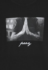 Black t-shirt featuring a black-and-white photo of hands in a praying position. Below, the text "pray" is printed in white script.
