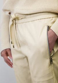 Ljusbeige joggerbyxor med dragsko i midjan, zippered sidoficka och slät tygtextur. Aningas med en krämfärgad stickad tröja.