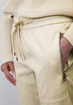 Ljusbeige joggerbyxor med dragsko i midjan, zippered sidoficka och slät tygtextur. Aningas med en krämfärgad stickad tröja.