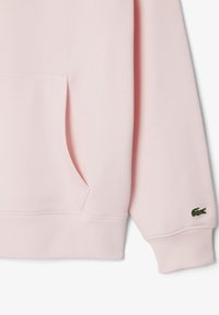 Růžová mikina s kapucí z měkké látky, s kapsou typu klokanka a žebrovanými manžetami. Na hrudi je malé zelené vyšívané logo.