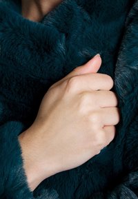 Vêtement en fausse fourrure teal avec une texture douce et moelleuse, présentant une coupe ample. Une main saisit le tissu, mettant en avant sa densité et sa chaleur.