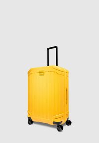Valigia rigida gialla con scanalature verticali, manico nero, quattro ruote per la mobilità e targa metallica con marchio sulla parte anteriore.