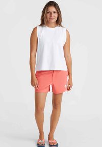 Vit ärmlös topp i kombination med korallfärgade shorts som har en logotyp. Modellen bär rosa sandaler med rosettaccenter. Enkel, avslappnad strandstil.