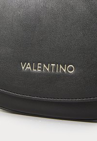 Svart teksturert skinnhåndveske med et gull "Valentino"-logo. Designet inkluderer subtil søm og en buet form.