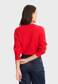 Maglione rosso a coste con un collo alto e spalle ribassate, abbinato a jeans in denim scuro a vita alta. Dettaglio visibile sulla schiena e maniche lunghe.