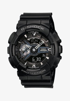 Črn G-Shock ura z teksturiranim gumijastim trakom, več kazalci, digitalnim prikazom in srebrnimi akcenti. Vsebuje oznake "Shock Resist" in "Casio WR20BAR".