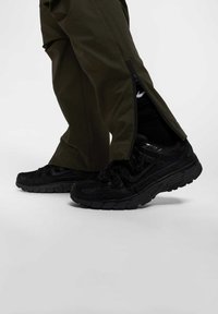 Sorte sneakers med teksturerede overflader og gummisåler, parret med olivengrønne bukser med lynlåse langs siderne.