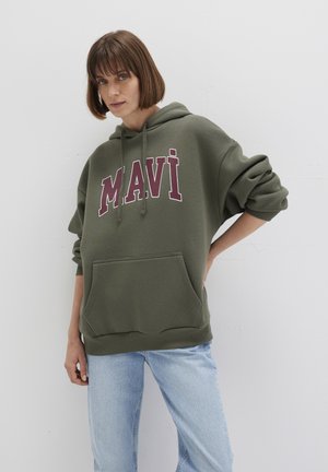 Olive green hoodie made of soft fabric, featuring a front pocket and bold maroon lettering "MAVI" on the chest. Paired with light blue jeans.