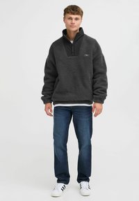 Fleece pullover in dark gray, featuring a black zip-up collar and chest panel, with textured fabric and logo detail. Worn with blue jeans.