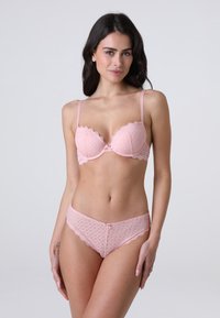 Donna con lunghi capelli scuri che indossa un reggiseno e delle mutandine in pizzo rosa chiaro coordinati, in piedi contro uno sfondo grigio chiaro uniforme.