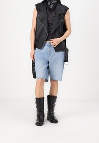 Černá kožená vesta bez rukávů přes puntíkovaný top, v kombinaci se světle modrými džínovými šortkami a černými kozačkami po kolena s detaily přezek.
