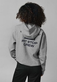 Sivá mikina s kapucňou s nápisom "PIT STOP CREW" vo námorníckej modrej farbe na chrbte. Mäkkučký materiál, voľný strih, so priestrannou kapucňou a dlhými rukávmi.