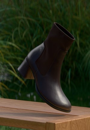 Dunkelbrauner Leder-Stiefelette mit Blockabsatz, die über einer Holzoberfläche mit grünem Laub im Hintergrund schwebt.