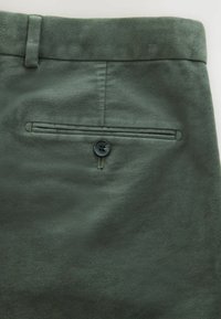 Dunkelgrüne maßgeschneiderte Hose mit glatter Textur, ausgestattet mit einer Gesäßtasche und einem einzelnen dunklen Knopfverschluss.
