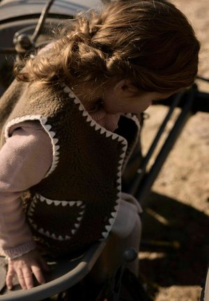 Barn som sitter på en benk utendørs, iført en brun vest med hvit kant over en rosa genser, ser nedover med krøllete hår bundet bak.