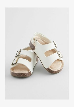 Sandalias sintéticas blancas con dos hebillas ajustables, paneles laterales recortados y una suela de corcho marrón texturizada para mayor tracción.