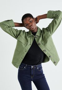 Olivengrønn denimjakke med krage, brystlommer og knappeåpning, kombinert med en svart topp og mørke jeans. Glatt tekstur.