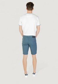 Camisa blanca de manga corta con ribetes negros, combinada con pantalones cortos azules y zapatillas blancas, vista desde atrás. Los pantalones cortos tienen bolsillos y cinturilla.