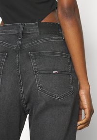 Jeans de mezclilla negra con un acabado texturizado, bolsillos traseros curvados y una etiqueta rectangular en la cintura que muestra un logo de la marca.