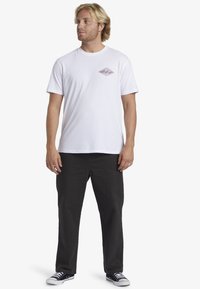 T-shirt bianco a maniche corte con una piccola grafica a diamante rossa e blu sul petto, abbinato a pantaloni neri e scarpe nere e bianche.
