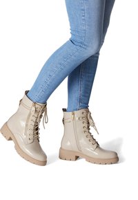 Beige patent leather lace-up boots with a chunky, textured rubber sole and gold hardware accents. Worn with light blue skinny jeans.