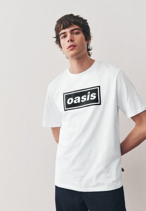 Mladý muž s krátkými hnědými vlasy, který má na sobě bílé tričko s černým ohraničeným nápisem „oasis“, stojí na jednoduchém světlém pozadí.