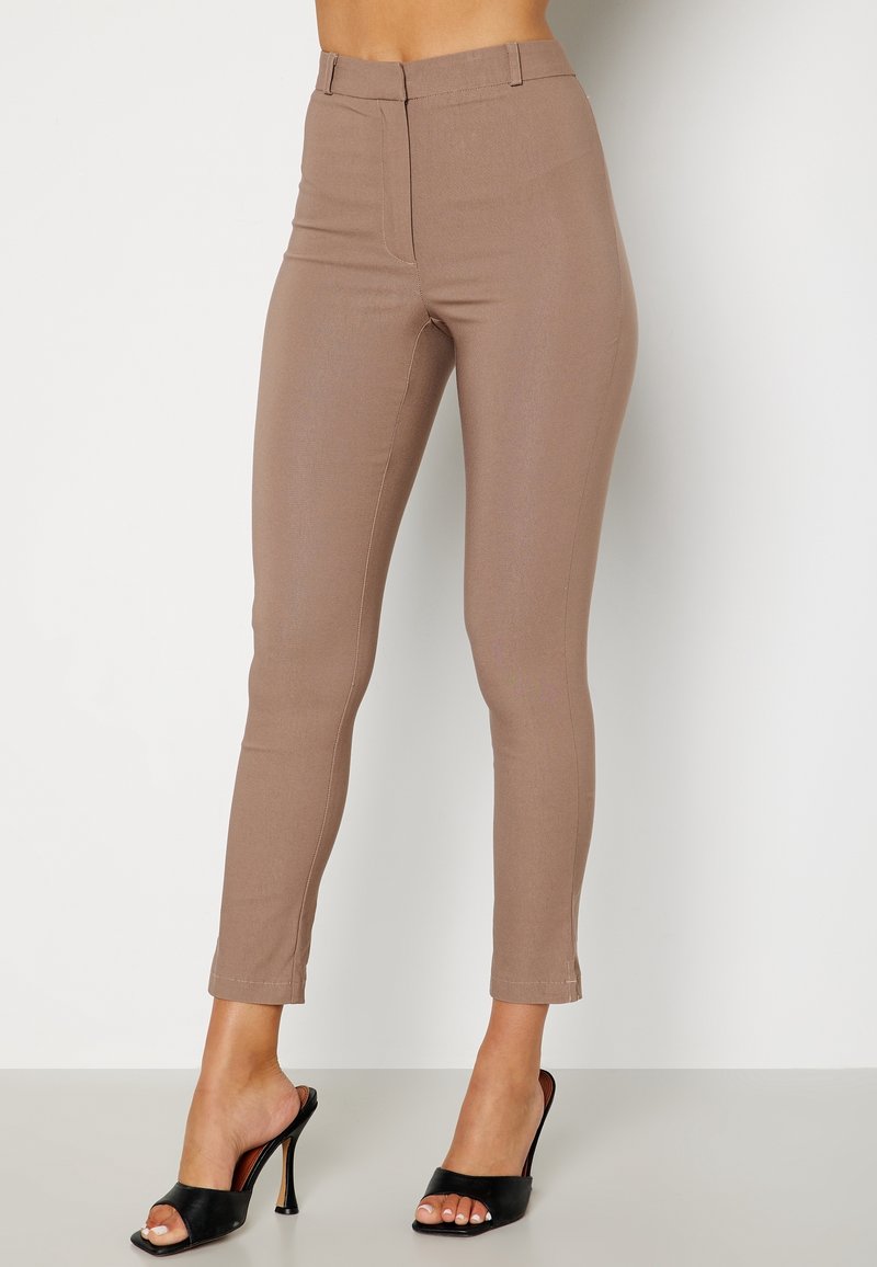 Femme portant un pantalon beige taille haute slim et des sandales à talons noires ouvertes, debout devant un fond clair uni.