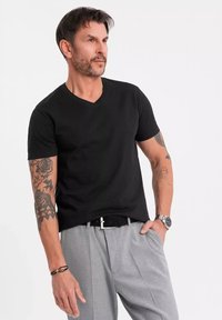 T-shirt noir à col en V en coton, associé à un pantalon beige clair sur mesure. Présente des tatouages sur les bras et une montre au poignet.