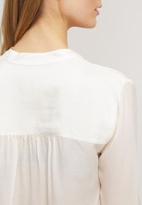 Žena s dlouhými rudými vlasy, oblečena v bílé blůze z texturované tkaniny s detailem a okrajem, zobrazená zezadu a z bočního profilu.