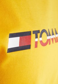 Žluté bavlněné tričko s výrazným nápisem "TOMMY" a designem loga v černé, bílé a červené. Hladká textura, volný střih.