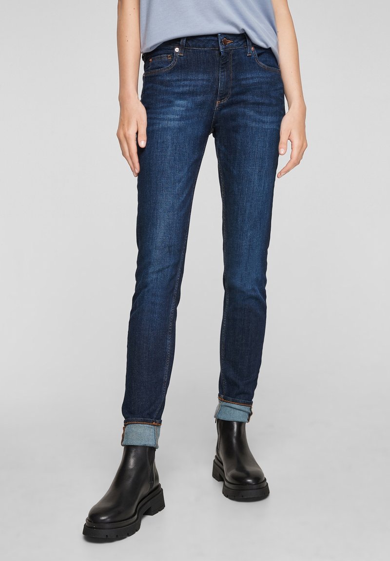 Dark blue skinny jeans with a slight fade, rolled cuffs, five-pocket design, and metal button fly, paired with black chunky boots.