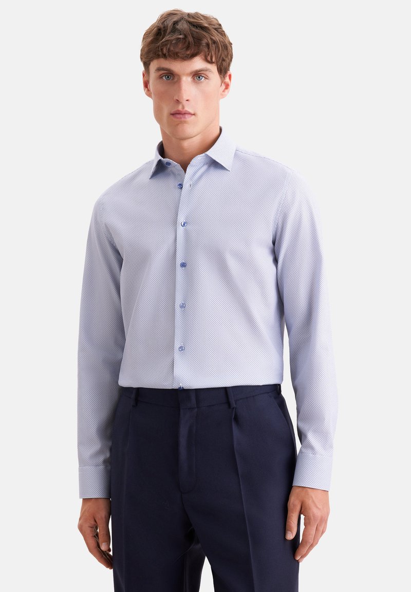 Homme portant une chemise à manches longues bleu clair à motifs, rentrée dans un pantalon de costume bleu marine foncé, debout devant un fond clair uni.