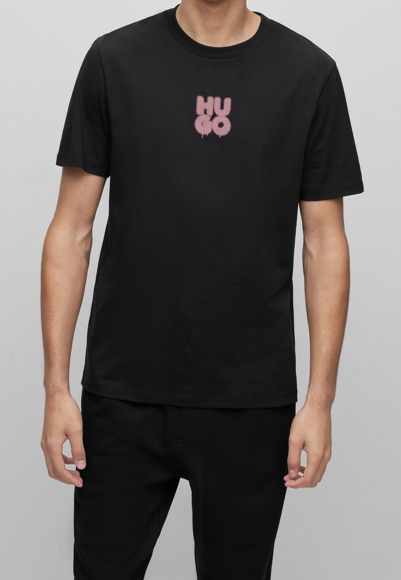 Sort bomulds t-shirt med et lyserødt spraymalet "HUGO" logo i midten. Korte ærmer, rund hals og standard pasform. Minimalistisk design.