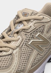 Κοντινή λήψη μπεζ αθλητικού παπουτσιού New Balance με υφή από δίχτυ, διακοσμητικά σχέδια, παχιά κορδόνια και το εμπορικό σήμα "N" στο πλάι.