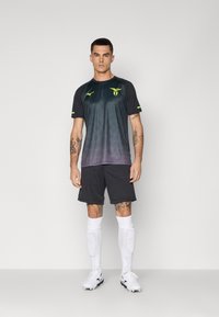 Mannlig idrettsutøver som står, iført svart og lilla gradient sportsdrakt, svarte shorts, hvite knelange sokker med leggskinn, og hvite fotballsko.