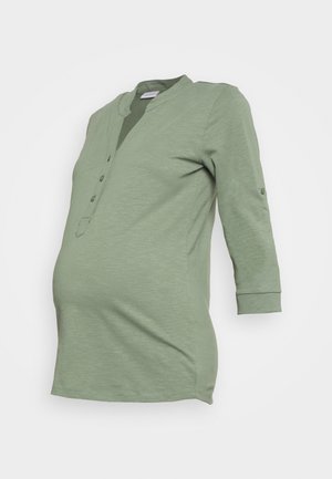 Světle zelený těhotenský top s tříčtvrtečními rukávy, zapínáním na knoflíky a přiléhavým pasem, navržený tak, aby se přizpůsobil rostoucímu bříšku.