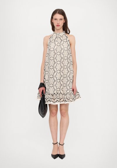Spitzen-Halterkleid in Creme und Schwarz, mit kreisförmigen Mustern und schwingendem Saum. Kombiniert mit einer schwarzen Clutch und spitzen Schuhen.