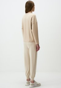 Felpa beige in cotone con spalle scese, abbinata a joggers beige con polsini elastici. Modello che indossa sneakers chiare.