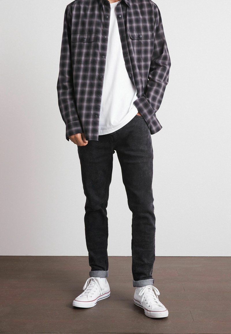 Camisa de franela a cuadros en negro y gris, camiseta de cuello redondo blanca, jeans oscuros, zapatillas blancas con detalles rojos, exhibidas contra un fondo neutro.