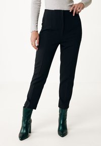 Höga svarta byxor med veck, i kombination med blanka mörkgröna ankelstövlar. Slätt tyg med skräddarsydd passform och kort längd.