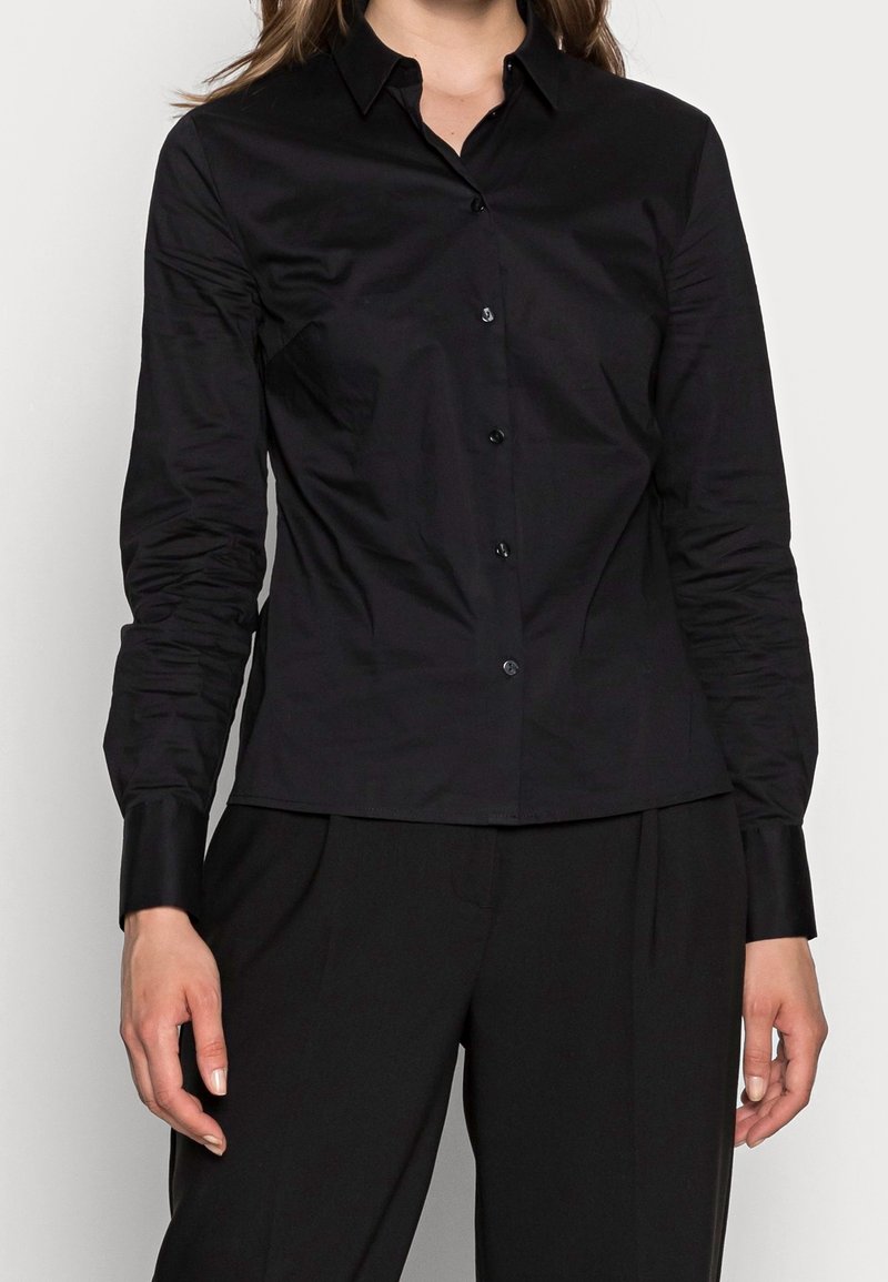 Femme portant une chemise noire cintrée à boutons avec manches longues et un pantalon noir, debout devant un fond clair uni.