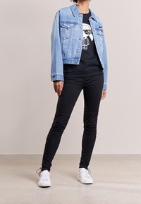 Persona che indossa una giacca di denim azzurro chiaro sopra una felpa nera con grafiche, pantaloni skinny neri e sneakers bianche, in piedi all'interno.