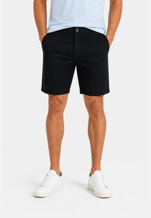 Mann mit schwarzen knielangen Shorts, weißen Sneakers und einem hellblauen Hemd mit den Händen in den Taschen vor einfachem Hintergrund.