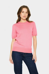 Kurzärmliger Rundhals-Pullover aus leichtem rosa Strick. Hat eine feine Textur mit gerippten Bündchen und Saum. Kombiniert mit dunkelblauen Jeans.