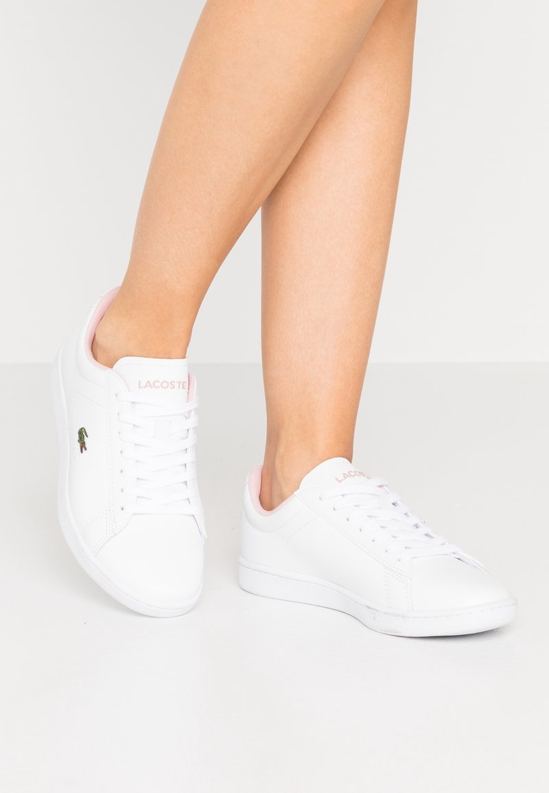Sneakers bianchi Lacoste con dettagli rosa indossati su una gamba contro uno sfondo semplice, che mettono in risalto il logo del marchio sul lato.